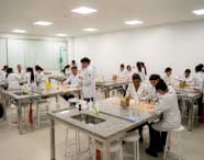 Visão ampla de um laboratório de Biomedicina com diversos alunos em jalecos brancos e luvas, realizando experimentos e atividades práticas nas bancadas equipadas com pias.
