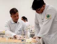 Professor de Biomedicina em jaleco branco e luvas orientando um aluno em uma aula prática, ambos examinando tubos de ensaio e vidrarias em uma bancada de laboratório.