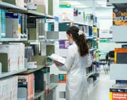 Aluna de Biomedicina em jaleco branco de costas, olhando e selecionando livros em uma estante da biblioteca universitária da Unoeste, focando em materiais de estudo.