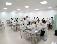 Vista panorâmica de um laboratório de Biomedicina com várias bancadas equipadas com pias, vidrarias e alunos em jalecos brancos realizando experimentos e aulas práticas.