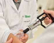 Aplicação de ventosa transparente no braço de estudante, que veste jaleco branco. Mão feminina segura bomba metálica preta para criar vácuo. No jaleco, logo "Unoeste" em verde. Fundo neutro destaca o foco no procedimento de ventosaterapia.
