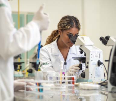 Perfil profissional Biomedicina , Jovem mulher de pele clara e cabelos longos e escuros, usando óculos e jaleco branco, sorri para a foto em ambiente interno com fundo claro. A iluminação destaca o rosto, transmitindo profissionalismo e contexto acadêmico no campus de Guarujá.
