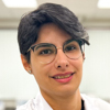 Renata, aluna do curso de biomedicina no campus Jaú, trajando jaleco branco. Ela está em uma sala de aula com fundo claro e paredes brancas, transmitindo um ambiente acadêmico organizado e propício ao aprendizado.