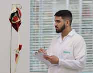 Faculdade de Biomedicina: Aluno de barba, com jaleco branco Unoeste, anota em uma prancheta enquanto observa um modelo anatômico de torso humano em um laboratório moderno, com persianas ao fundo, compondo um cenário de aula prática, limpo e organizado.