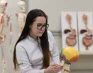Faculdade de Biomedicina: Aluna de cabelos escuros e óculos, com jaleco branco Unoeste, segura e observa um modelo anatômico de crânio e coluna vertebral em um laboratório moderno, com esqueletos ao fundo, compondo um cenário de aula prática, limpo e organizado.