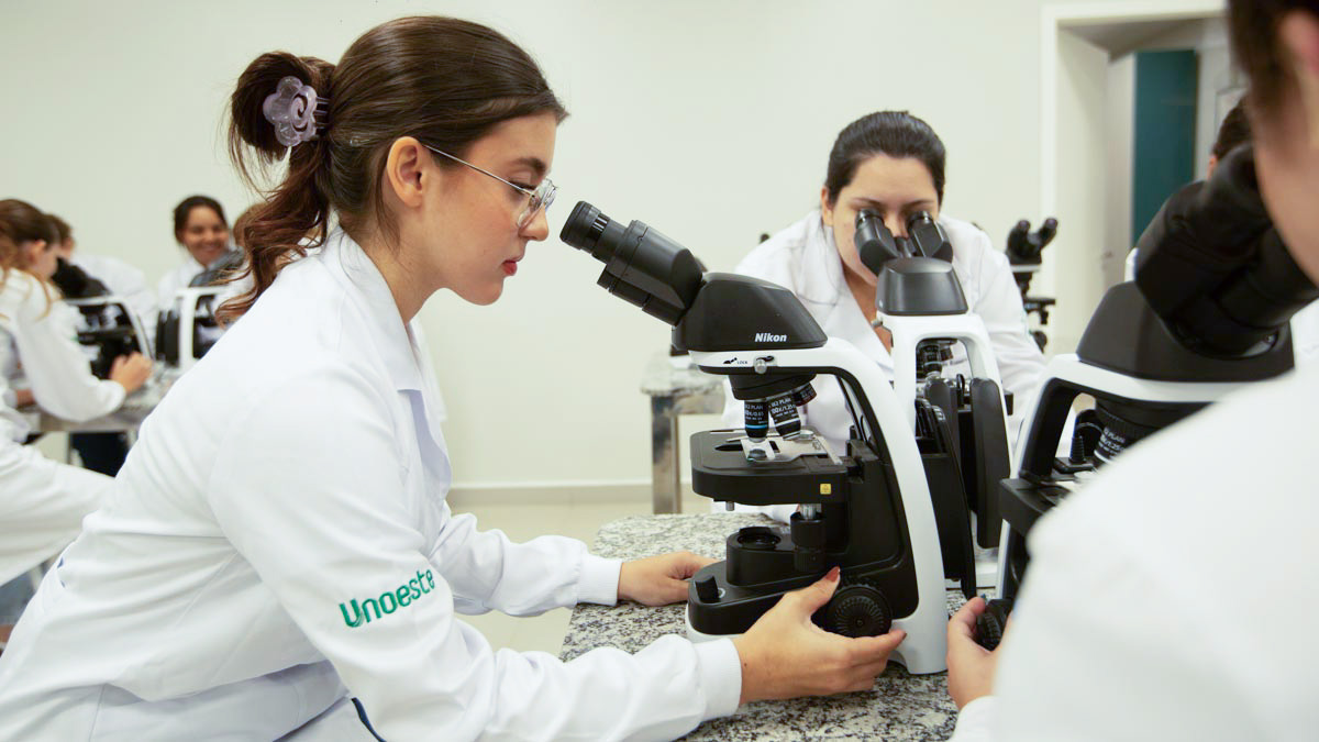 Aluna de Biomedicina em jaleco branco e óculos examinando uma amostra em um microscópio óptico durante uma aula prática em laboratório.