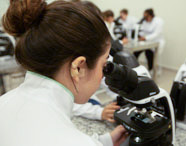Close-up de aluna de Biomedicina com cabelo preso observando uma amostra através de um microscópio óptico Nikon em uma aula de laboratório.