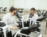 Duas alunas de Biomedicina em jalecos brancos concentradas examinando amostras em microscópios ópticos durante uma aula prática no laboratório.
