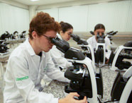 Aluno de Biomedicina em jaleco branco e óculos utilizando um microscópio óptico Nikon em aula prática de laboratório, com outros alunos ao fundo.