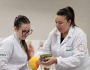 Faculdade de Biomedicina: Duas alunas com jalecos brancos Unoeste, uma delas com óculos, observam e interagem com um modelo anatômico de crânio e coluna vertebral em um laboratório moderno, compondo um cenário de aula prática, limpo e organizado.