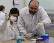 Faculdade de Biomedicina: Professor e aluna com jalecos brancos e luvas, manuseiam tubos de ensaio e pipetas com líquidos coloridos em uma bancada de laboratório moderno, com equipamentos de vidro ao fundo, compondo um cenário de aula prática, limpo e organizado.