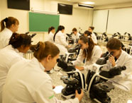 Laboratório de Biomedicina bem equipado, com diversos alunos em jalecos brancos utilizando microscópios ópticos em suas bancadas, demonstrando uma aula prática.