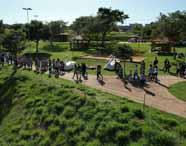 Turma do curso de Ciências Biológicas, formação pedagógica, da Unoeste realiza atividade em campo em um parque arborizado. Os estudantes caminham por uma trilha pavimentada, cercados por gramado verde, árvores e esculturas brancas de cisnes. O cenário é aberto, com quiosques ao fundo e clima ensolarado.