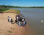 Grupo de alunos do curso de Ciências Biológicas, formação pedagógica, da Unoeste participa de atividade externa à beira de um lago. Os estudantes estão reunidos na areia próxima à água, observando o ambiente natural. Ao fundo, há vegetação, céu azul e algumas boias amarelas delimitando a área do lago.