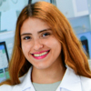 Imagem de Sara, aluna do curso de medicina no campus Presidente Prudente, vestindo jaleco branco. Ela está em um ambiente interno com equipamentos médicos e monitores ao fundo, indicando um espaço de aprendizado prático na área médica. A iluminação clara contribui para destacar os detalhes do local.