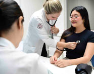 Em ambiente clínico, estudante de Medicina realiza exame físico em paciente, utilizando estetoscópio para auscultar o tórax. Outra aluna observa atentamente. As duas estão de jaleco branco, enquanto a paciente veste camiseta preta e está sentada à mesa.