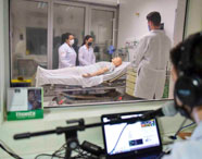 Estudantes de Medicina realizam simulação de atendimento hospitalar em laboratório. Um grupo observa e executa procedimentos em manequim adulto deitado em maca, enquanto instrutor monitora a atividade através de vidro, usando microfone e fones de ouvido.