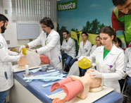 Alunos de Medicina treinam técnicas de ventilação manual em manequins de simulação, utilizando ambus de diferentes cores. A atividade ocorre em sala equipada, com materiais médicos sobre a mesa e outros estudantes assistindo, todos de jaleco branco.