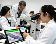 Sala de microscopia com estudantes de Medicina analisando lâminas biológicas. Uma aluna observa pelo microscópio, outra manuseia tablet com imagem ampliada de tecido. Professor acompanha a atividade, todos usando jaleco branco em ambiente moderno.