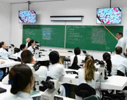 Aula teórica de histologia no curso de Medicina, com estudantes atentos em sala equipada com microscópios individuais. Professor explica conteúdo no quadro verde, apontando desenho esquemático de tecido, enquanto imagens ampliadas são projetadas nas telas.