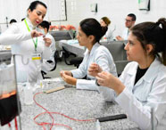 Estudantes de Medicina praticam técnicas de preparo e administração de medicamentos em laboratório. Sentadas à bancada de granito, elas manuseiam seringas e frascos, sob orientação de professora identificada por crachá verde da Unoeste.