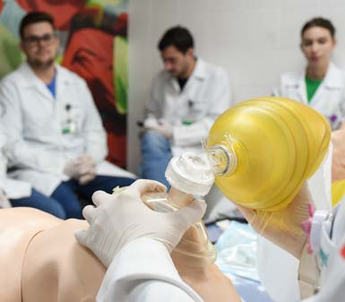 Estudantes do curso de Medicina da Unoeste em Presidente Prudente participam de treinamento prático. Em destaque, mãos com luvas realizam ventilação manual com ambu em manequim de simulação. Ao fundo, outros alunos observam sentados, todos usando jaleco branco em ambiente de laboratório.