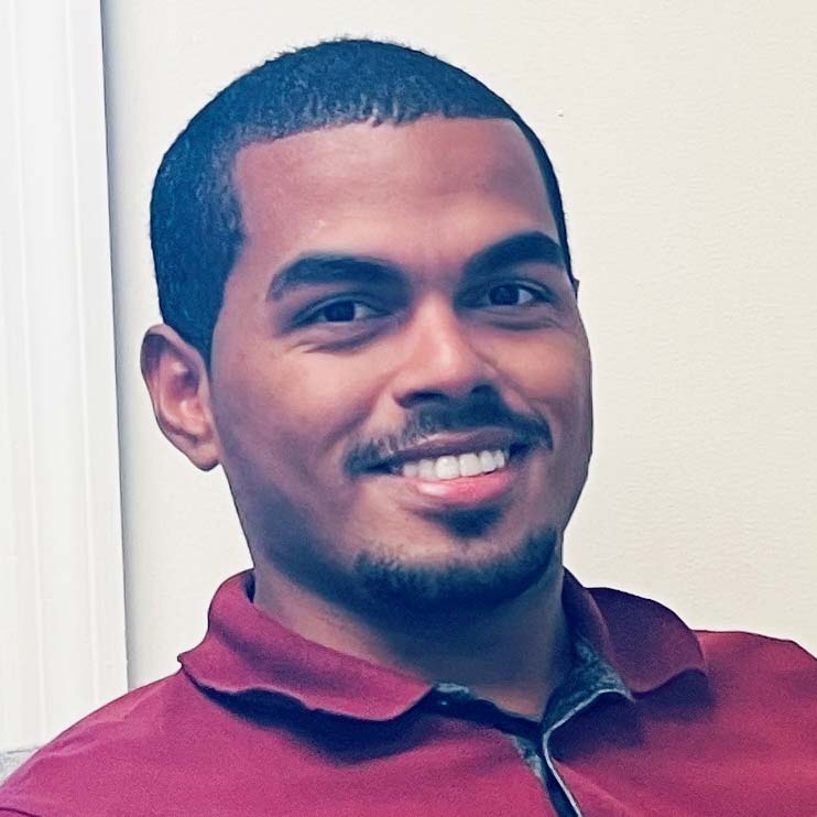 João Pedro, egresso de administração, posando em um ambiente interno com um fundo claro. Ele usa uma camisa polo vinho e tem barba rala, com cabelos curtos e um sorriso amigável.
