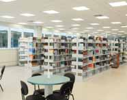 Faculdade de administração em Guarujá biblioteca sala de convivência prateleiras: imagem mostra prateleiras repletas de livros organizados e mesas redondas com cadeiras para estudo em grupo. O espaço é amplo, bem iluminado, moderno e pensado para estimular a convivência, pesquisa e aprendizado colaborativo entre os alunos do curso de administração.