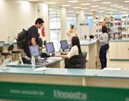 Faculdade de administração em Guarujá biblioteca: balcão de atendimento com computadores, funcionários auxiliando estudantes, prateleiras de livros ao fundo e ambiente amplo e iluminado. O espaço destaca o suporte ao estudante, a variedade de recursos e a importância do atendimento humanizado para uma experiência acadêmica completa.