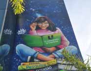 Faculdade de administração em Guarujá grafite fachada: painel artístico na lateral do prédio mostra uma jovem sorrindo, lendo um livro verde com a frase “O conhecimento te transforma”, sobre fundo estrelado.