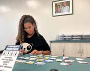 Faculdade de administração em Guarujá jogo interativo com alunos: estudante participa de dinâmica no balcão de atendimento, com cartões coloridos organizados sobre a mesa. O ambiente é acolhedor, com sinalização de atendimento prioritário e decoração leve, reforçando o apoio ao aluno, acessibilidade e incentivo a metodologias participativas no ensino superior.