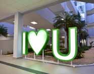 Faculdade de administração em Guarujá letreiro I love Unoeste: letreiro iluminado “I coração U” em destaque no hall interno, entre jardins e palmeiras. O espaço moderno, com design convidativo e paisagismo, reforça o orgulho institucional e o ambiente acolhedor do campus, promovendo integração, pertencimento e identidade universitária.