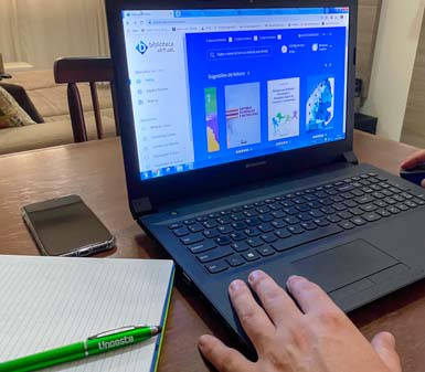 Acesso à biblioteca digital Unoeste, a imagem mostra uma pessoa usando um notebook sobre uma mesa de madeira, navegando por uma plataforma de biblioteca digital. Ao lado do computador há um caderno aberto, uma caneta verde com a inscrição Unoeste e um smartphone desligado. O ambiente é residencial e acolhedor, com sofá ao fundo, indicando rotina de estudos flexível. A cena destaca a praticidade do acesso remoto a conteúdos acadêmicos e a integração de recursos físicos e digitais no aprendizado.