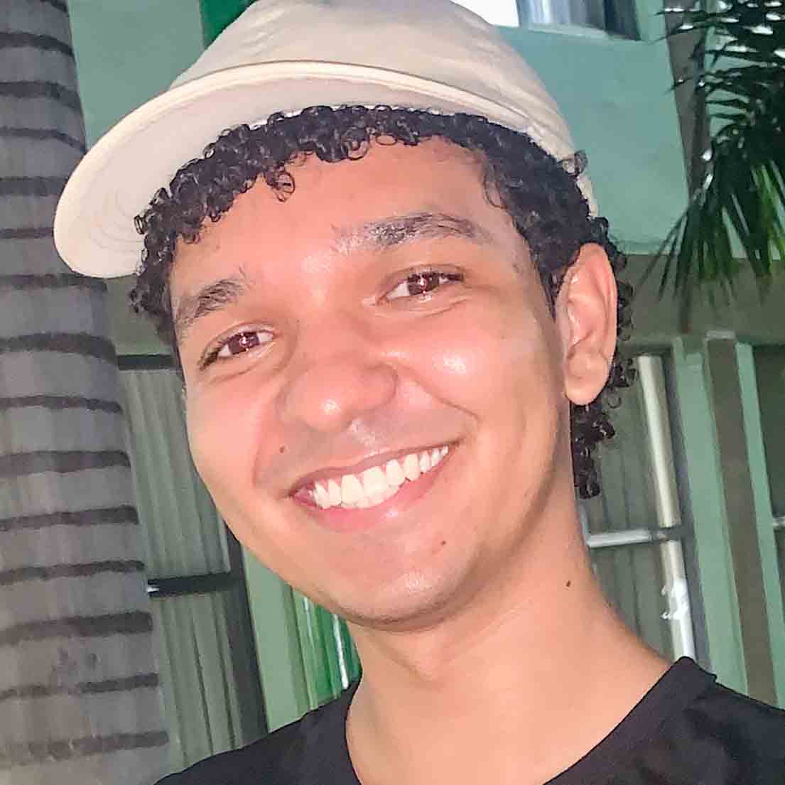 João Pedro, aluno de administração, posando em um ambiente externo com um fundo verde. Ele usa um boné bege e uma camiseta preta, com cabelos curtos e cacheados e um sorriso largo.