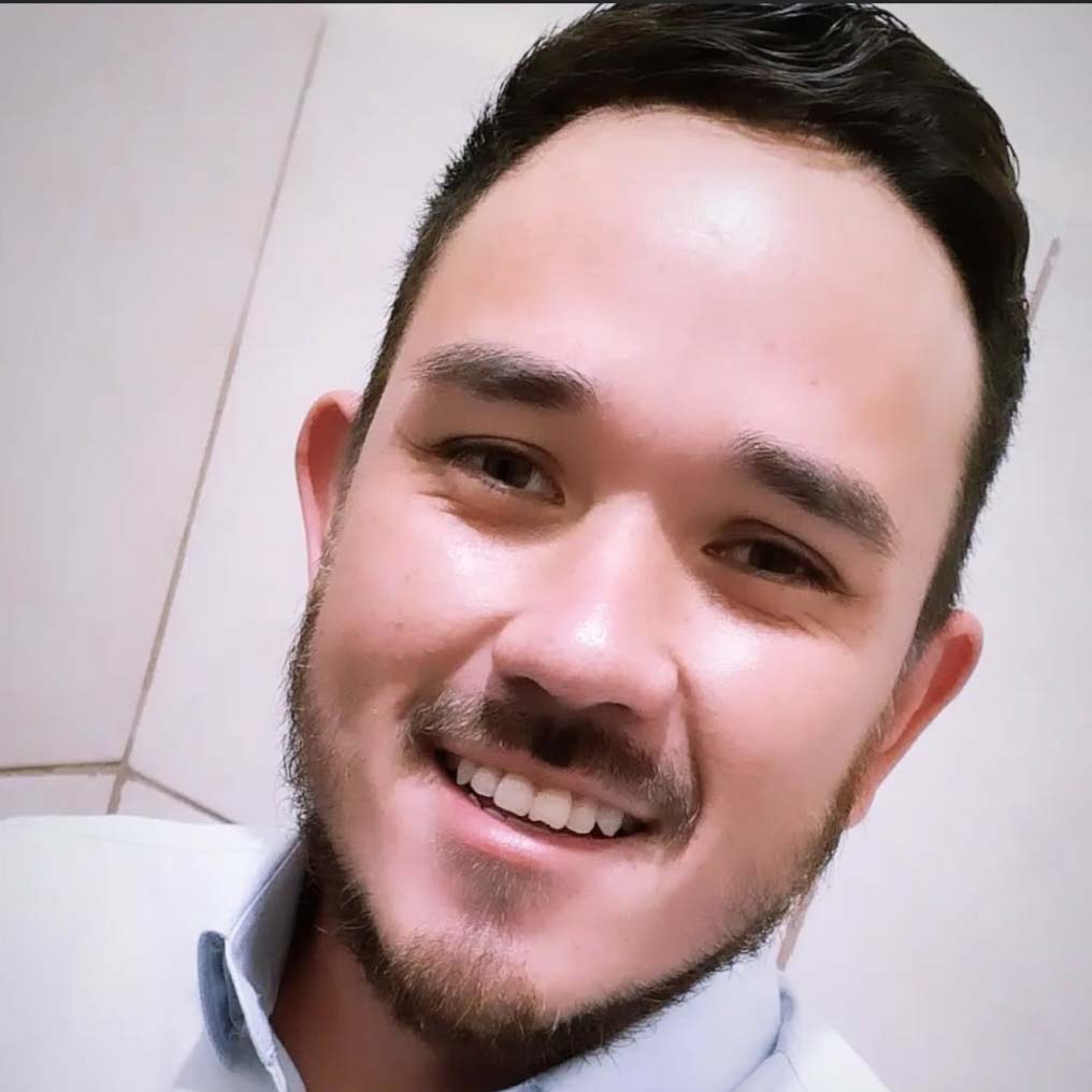 Murilo, aluno de administração, posando em um ambiente interno com um fundo claro. Ele usa uma camisa social azul clara e tem barba rala, com cabelos escuros e um sorriso largo.