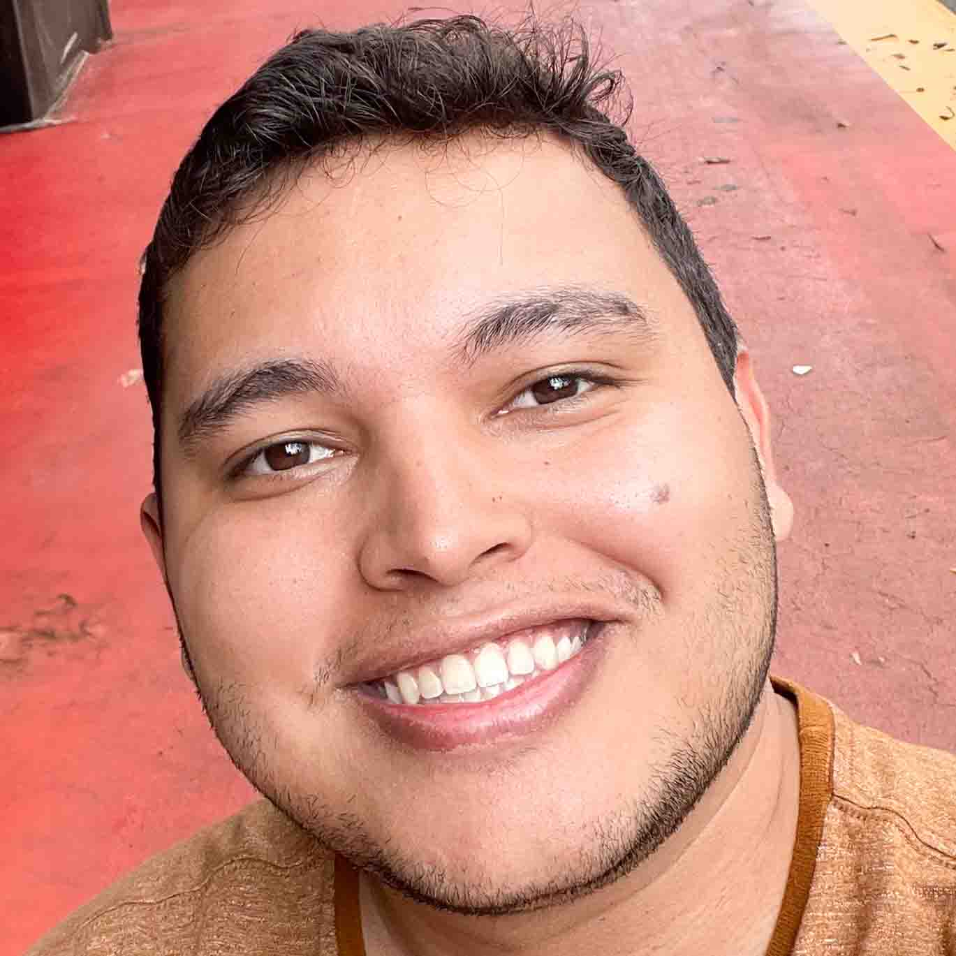 Paulo, aluno de administração, posando em um ambiente externo com um piso vermelho ao fundo. Ele usa uma camiseta marrom e tem barba rala, com cabelos escuros e um sorriso largo.