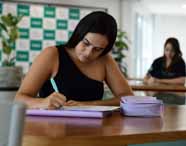 Faculdade de administração em Jaú, aluna concentrada faz anotações em mesa de estudo, utilizando estojo e caderno, enquanto ao fundo outra estudante também se dedica às atividades acadêmicas. O ambiente interno, bem iluminado e com plantas, reforça o clima de foco, incentivo ao aprendizado e dedicação dos alunos do curso de administração.
