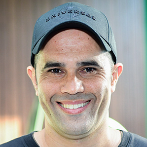 Steve, aluno do curso de direito, trajando uma camiseta preta e um boné cinza. Ele está em um ambiente externo com fundo composto por madeira e detalhes verdes, sugerindo um espaço institucional. A iluminação natural destaca os elementos da vestimenta e cria um contraste suave com o cenário ao redor, transmitindo um contexto casual e acadêmico.