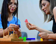 Duas pessoas interagem em uma mesa utilizando jogos pedagógicos e peças coloridas, como blocos de encaixe e base de madeira. A atividade representa práticas lúdicas aplicadas ao ensino de Psicologia, promovendo o desenvolvimento cognitivo e social em ambiente universitário.