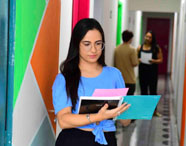 Aluna caminha por corredor colorido com paredes decoradas em tons de azul, verde e laranja, segurando cadernos e folhas. Ao fundo, outros estudantes conversam próximos às portas das salas. O ambiente transmite dinamismo e acolhimento no cotidiano acadêmico do curso de Psicologia.