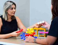 Duas pessoas utilizam brinquedos educativos de montar, com bolas plásticas coloridas conectáveis, sobre uma mesa. A cena evidencia atividades lúdicas e pedagógicas voltadas ao desenvolvimento infantil, frequentemente utilizadas nas práticas do curso de Psicologia.