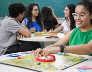 Sala de aula com grupos de estudantes jogando jogos de tabuleiro e atividades lúdicas em mesas redondas. Ao fundo, quadro verde. Os jogos estimulam habilidades cognitivas e sociais, integrando teoria e prática no curso de Psicologia.