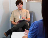 Estudante de Psicologia realiza atendimento individual em clínica-escola, sentado em poltrona com prancheta e crachá da instituição Unoeste. Em primeiro plano, paciente de costas. Ambiente claro e confortável, ideal para sessões supervisionadas de psicoterapia.