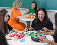 Grupo de estudantes em sala de aula participando de atividades com jogos de tabuleiro, incluindo jogos de perguntas e adivinhação. Ao fundo, quadro verde e professora acompanhando. Atividades desenvolvem raciocínio, memória e interação entre alunos do curso de Psicologia.