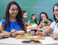 Estudantes de Psicologia manipulam jogo de madeira educativo em sala de aula, focados na atividade. Ao fundo, outros grupos também participam de dinâmicas lúdicas. O uso de jogos pedagógicos auxilia no desenvolvimento de competências cognitivas e emocionais.