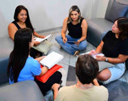 Grupo de cinco pessoas sentadas em círculo no chão, em espaço com sofás cinza, trocando ideias e fazendo anotações. Ambiente descontraído e colaborativo, típico de dinâmicas em grupo e rodas de conversa do curso de Psicologia.