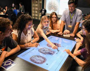 Turma de Psicologia reunida em laboratório tecnológico, em torno de mesa digital sensível ao toque, analisando imagens anatômicas do cérebro. Professor acompanha o grupo, projetor exibe imagem cerebral ao fundo. O ambiente destaca o uso de tecnologia avançada no ensino universitár