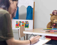 Ambiente infantil decorado para atividades lúdicas, com mesa pequena, livros abertos, cadeira infantil e grande urso de pelúcia usando boina. Ao fundo, painel colorido com desenhos de trem e cortinas azuis. Espaço voltado para práticas de Psicologia escolar e atendimento infantil.