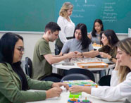 Sala de aula do curso de Psicologia com estudantes em mesas redondas, manuseando jogos pedagógicos e caixas de atividades. Professora acompanha os grupos, quadro verde ao fundo com tópicos escritos. O ambiente favorece aprendizagem ativa, interação e aplicação prática dos conteúdos.