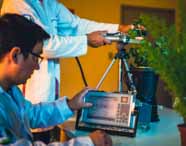 Dois estudantes do curso de Ciências Biológicas da Unoeste desenvolvem atividade prática em laboratório. Um deles opera um aparelho eletrônico com tela exibindo gráficos, enquanto o outro manuseia sensor conectado a plantas em vasos. O cenário destaca o uso de tecnologia aplicada ao estudo das ciências biológicas.